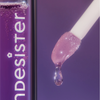 Eine Nahaufnahme des Applikators des BLONDESISTER 3 IN 1 CUTE LIPPY LIP OIL, von dem violetter Lipgloss herabtropft, neben der violetten Tube.
