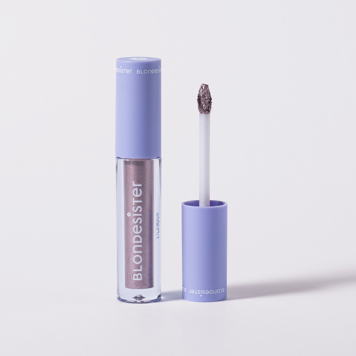 Eine Tube BLONDESISTER GLOWEYE EYESHADOW mit einer hellvioletten Kappe und dem dazugehörigen Applikator, der daneben liegt.
