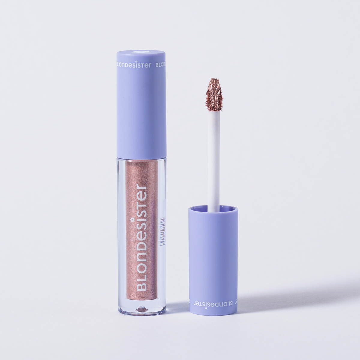 Ein geöffneter BLONDESISTER GLOWEYE EYESHADOW gibt den Blick auf seinen Applikator frei und enthüllt unter einer lavendelfarbenen Kappe schimmerndes rosa Pigment.
