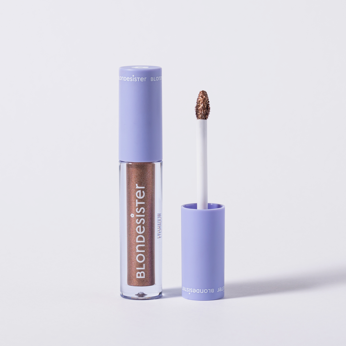 Eine Tube BLONDESISTER GLOWEYE EYESHADOW mit violetter Kappe, abgebildet in geöffnetem Zustand, mit schimmerndem Braun auf dem Applikator.