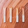 Vier Tuben Catrice Cosmetics Under Eye Brightener Liquid in verschiedenen Farbtönen sind auf einem strukturierten pfirsichfarbenen Hintergrund abgebildet.