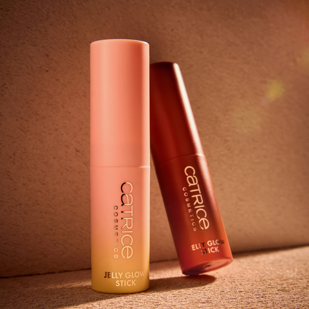 Zwei Catrice Cosmetics MIDNIGHT SUN Jelly Glow Sticks, einer in Pfirsich und einer in Bronze, werden auf einer strukturierten Oberfläche unter warmem Licht präsentiert.