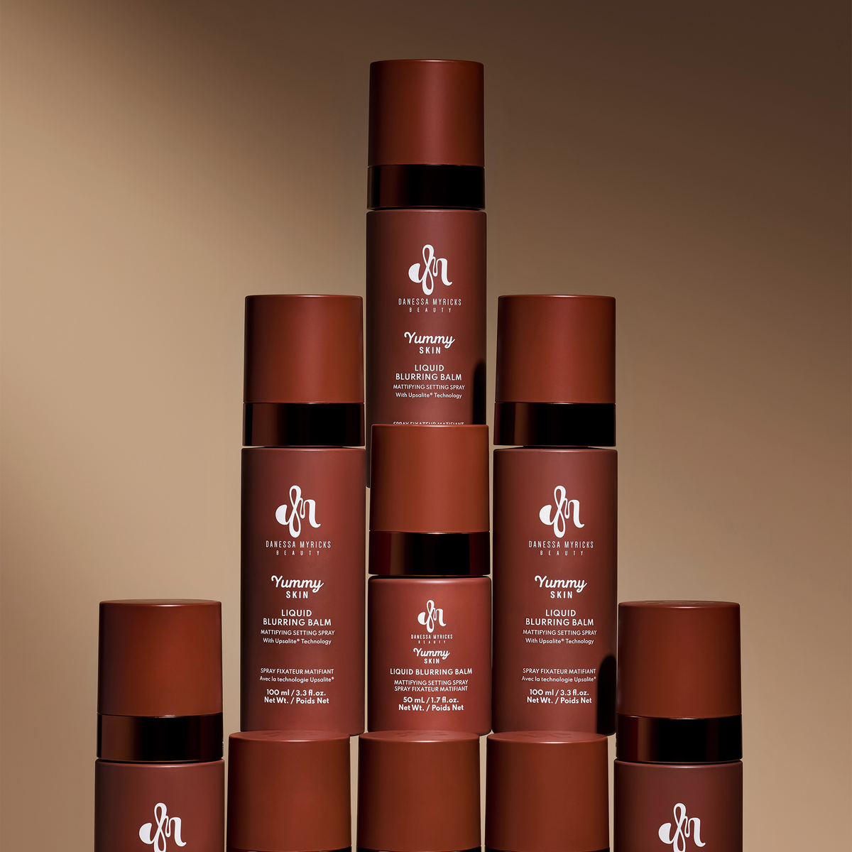 Mehrere braune Flaschen Danessa Myricks Yummy Skin Liquid Blurring Balm Spray in 50 ml und 100 ml sind pyramidenförmig vor braunem Hintergrund angeordnet.