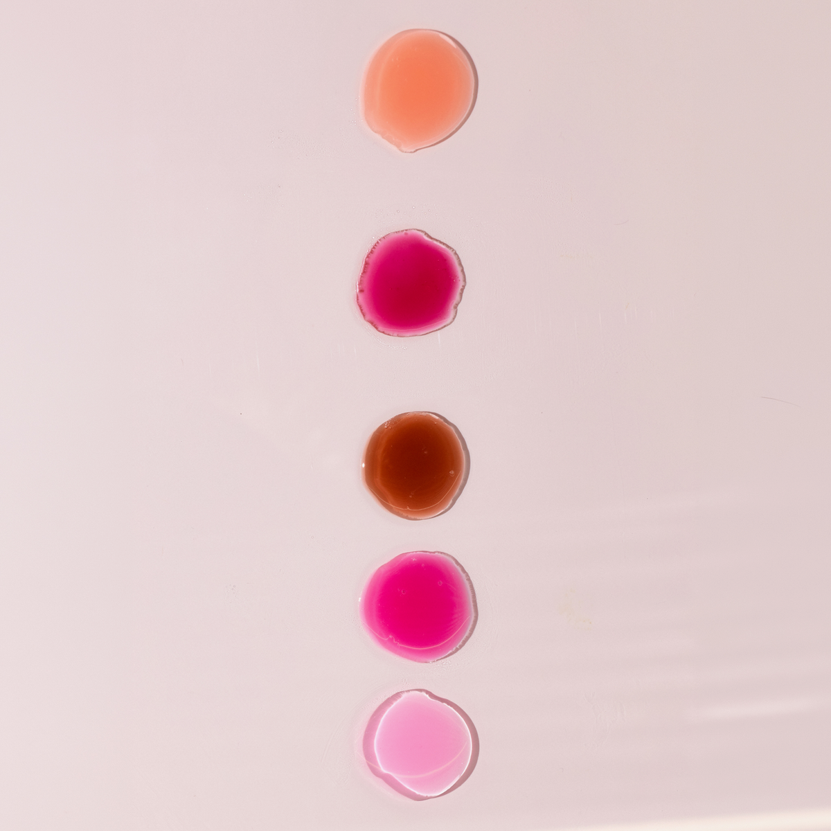 Fünf Farbnuancen des Dominique Cosmetics Juicy Lip Balm vertikal auf einer hellrosa Fläche aufgetragen.