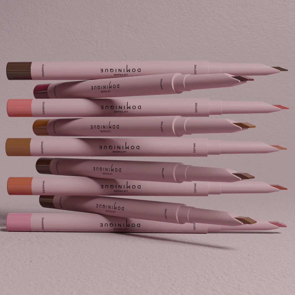 Ein Stapel Dominique Cosmetics Lip Frame Pencils in verschiedenen Farbtönen, horizontal angeordnet vor einem hellrosa Hintergrund.