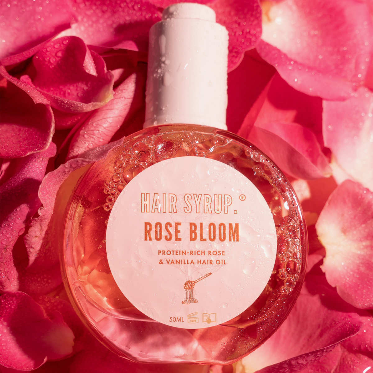 Eine Flasche Hair Syrup Rose Bloom Öl ist von rosa Rosenblättern umgeben.