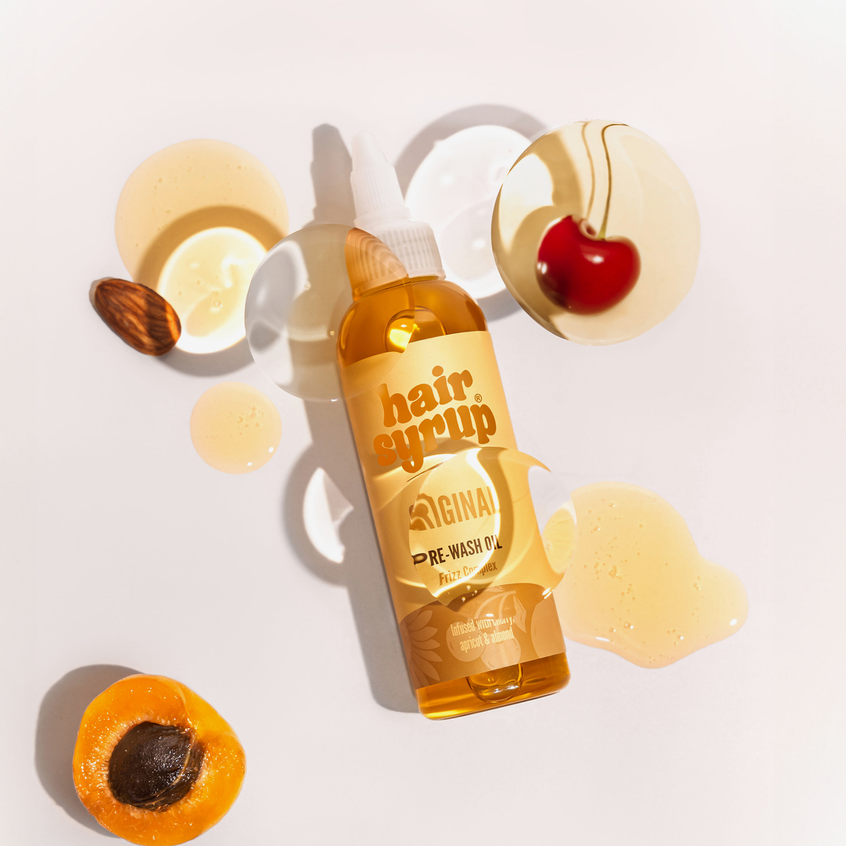 Eine Flasche Hair Syrup's The Original Pre-wash Oil, umgeben von verschüttetem Öl, einer Kirsche, einer Mandel und einer halbierten Aprikose.