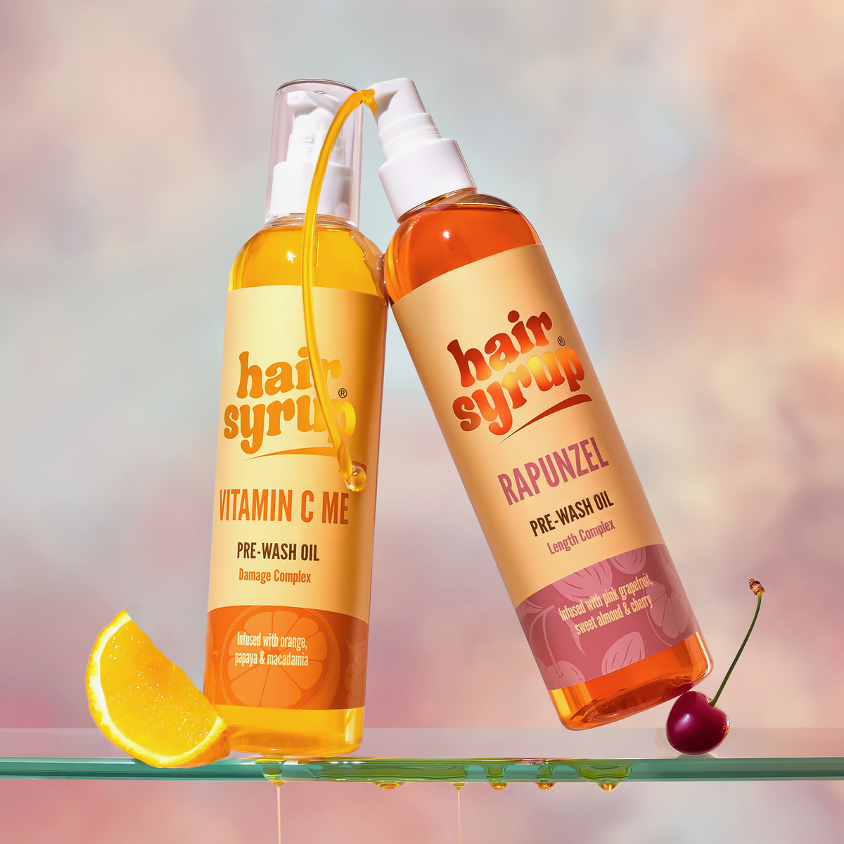 Zwei Flaschen Pre-Wash-Öls von Hair Syrup mit einer Orangenscheibe und einer Kirsche auf einem Glasregal.