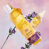 Auf einem pastellfarbenen Hintergrund sind zwei Flaschen des Vorwaschöls Hair Syrup Zen mit honigartiger Textur und Lavendelblüten abgebildet.