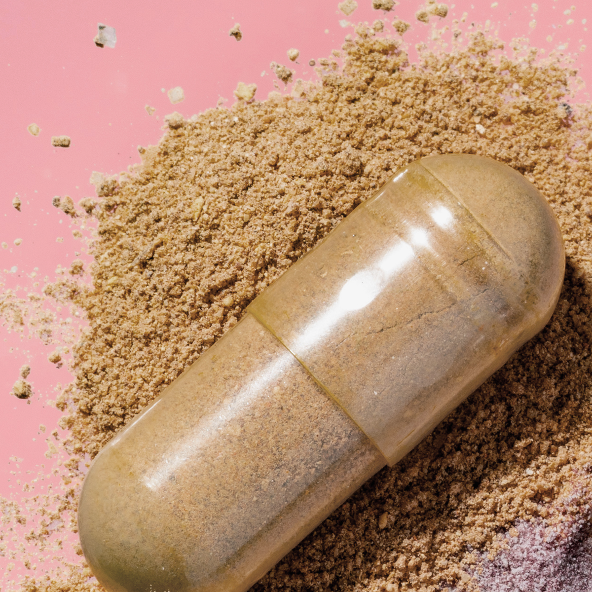 Transparente vegane Kapsel mit beigem Pulver auf rosafarbenem Hintergrund, umgeben von aufgestreutem Pflanzenpulver.