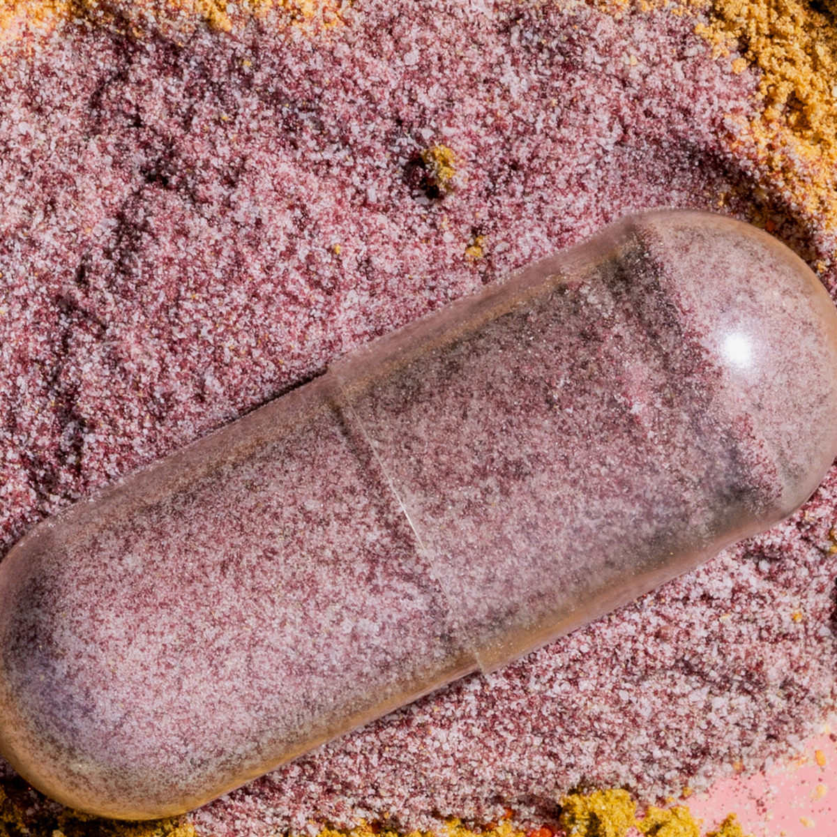 Nahaufnahme einer transparenten Kapsel auf rosafarbenem Pulver mit gelblichen Akzenten – symbolische Darstellung eines Nahrungsergänzungsmittels.