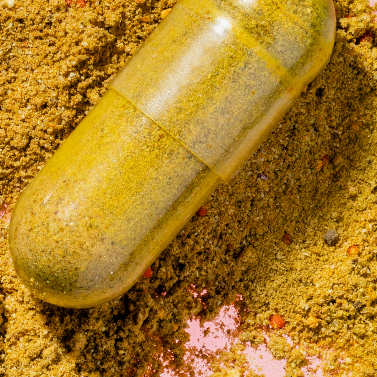 Transparente Kapsel mit gelblich-grünem Pulver auf körnigem Untergrund, Detailaufnahme eines Nahrungsergänzungsmittels.