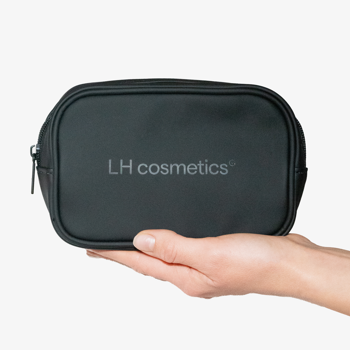 Vor einem schlichten Hintergrund hält eine Hand „The little black bag“ von LH Cosmetics – eine elegante schwarze Tasche mit weißem Logo.