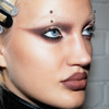 Eine Frau trägt ein auffälliges Make-up mit The Single von LH Cosmetics, Augenbrauenpiercings und Haarspangen vor einer weißen Backsteinwand.
