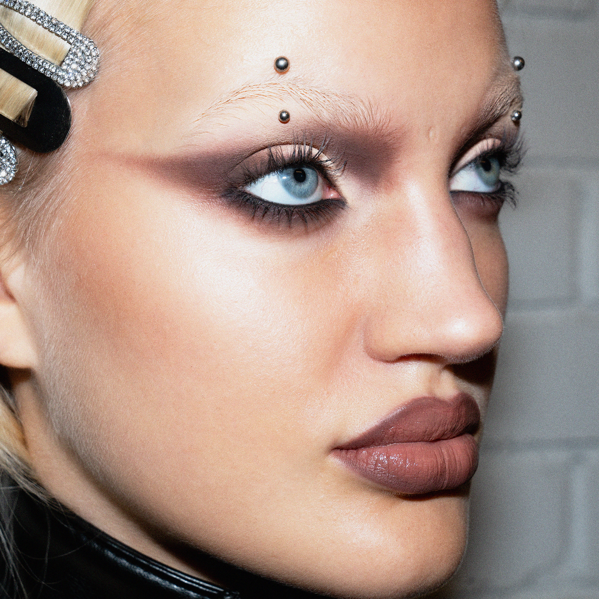 Eine Frau trägt ein auffälliges Make-up mit The Single von LH Cosmetics, Augenbrauenpiercings und Haarspangen vor einer weißen Backsteinwand.