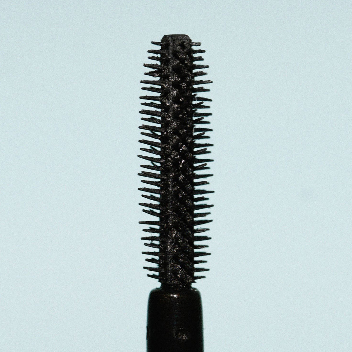 Nahaufnahme einer schwarzen Mascara-Bürste mit kurzen, dichten Gummiborsten vor hellblauem Hintergrund.
