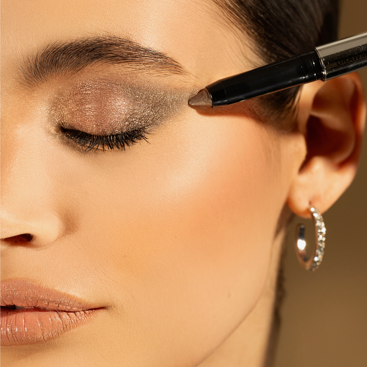 Nahaufnahme eines geschlossenen Auges mit schimmerndem braun-grauem Lidschatten, aufgetragen mit einem braunen Lidschattenstift am äußeren Augenwinkel.