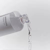 Transparente Flasche des Mixsoon Heartleaf Toners, aus der klarer Toner fließt, vor neutralem Hintergrund fotografiert.
