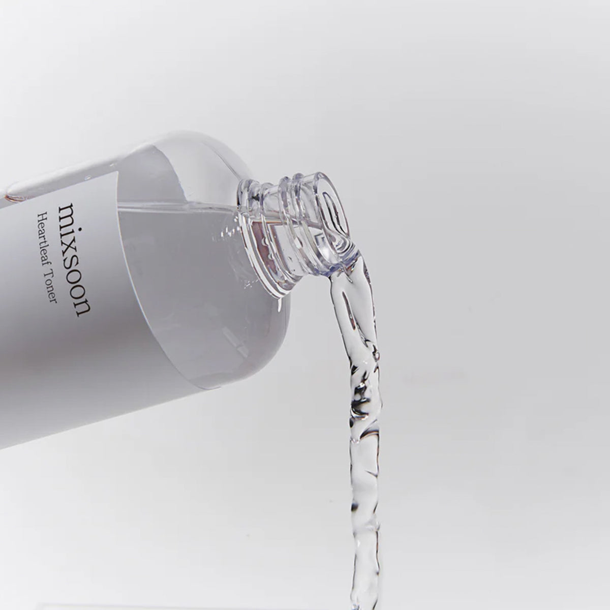 Transparente Flasche des Mixsoon Heartleaf Toners, aus der klarer Toner fließt, vor neutralem Hintergrund fotografiert.