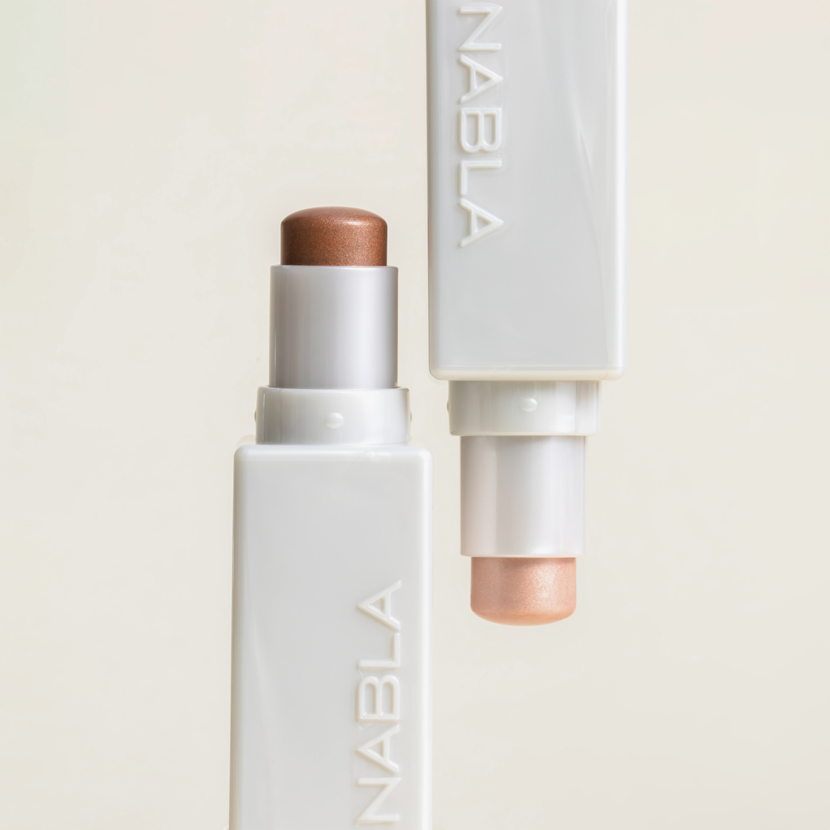 Zwei „Art Of Skin Glazing Sticks“ von NABLA Cosmetics in den Farben Bronze und Champagner stehen aufrecht in weißen Tuben vor einem hellen Hintergrund.