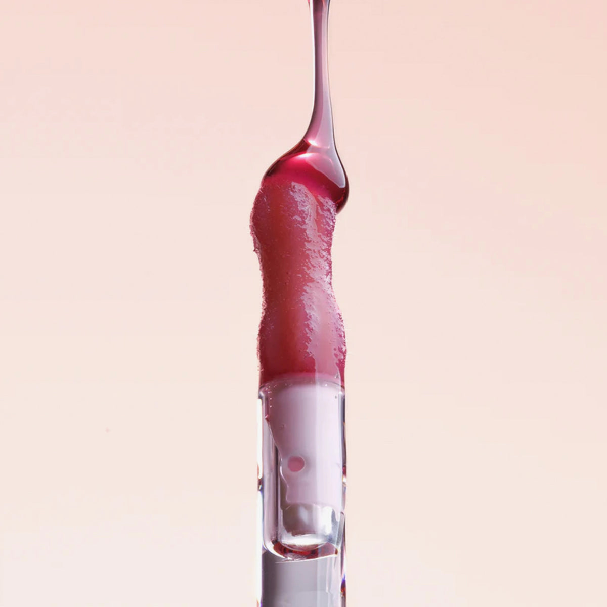Ein rötlicher Tropfen Lip Candy Oil Black Cherry gleitet vor einem hellrosa Hintergrund über den Applikator. 