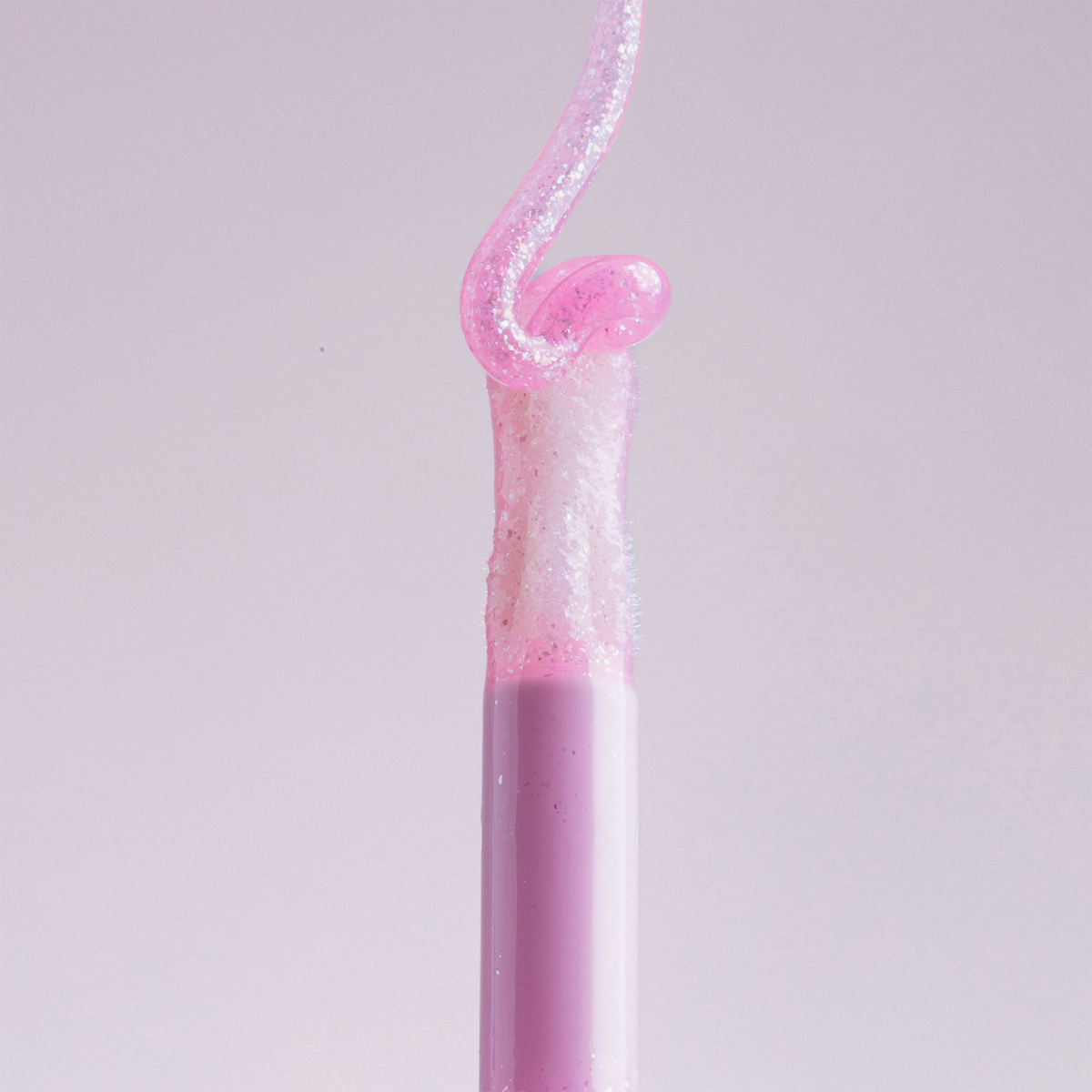 Aus dem „Lip Candy Oil“ von NABLA Cosmetics erhebt sich ein rosafarbener, glänzender Wirbel, der mit seiner eleganten rosa Tube vor einem blassen Hintergrund harmoniert.