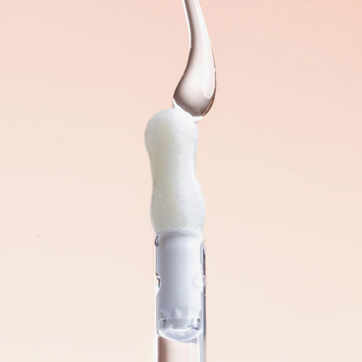 Ein transparenter Tropfen Lip Candy Oil Salted Caramel gleitet vor einem hellbeigenr Hintergrund über den Applikator. 