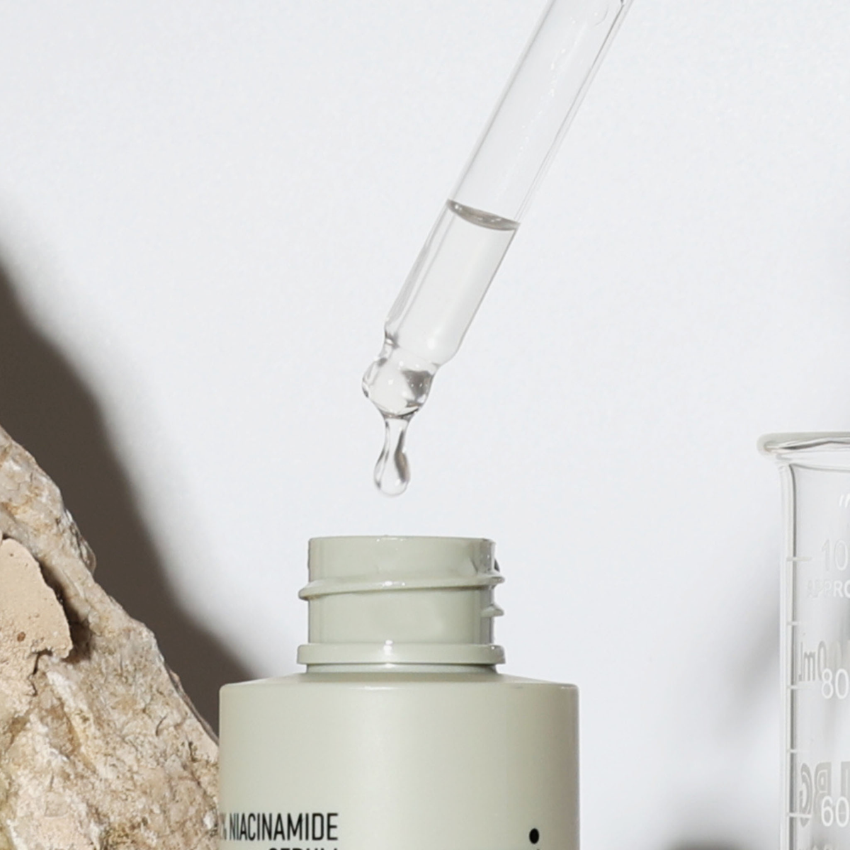 Ein Tropfer mit klarer Flüssigkeit schwebt über einer offenen NERDS. 12% Niacinamid + 1% Zink-PCA-Serum-Flasche, die neben einem Stein und einem Glasbecher steht.