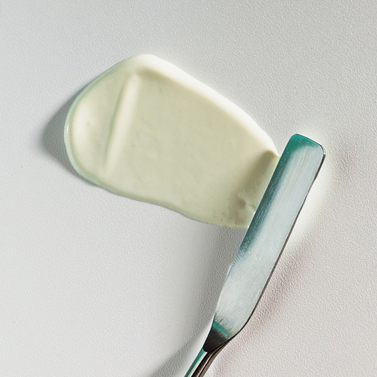 Ein Metallmesser verteilt eine gleichmäßige Schicht NERDS. Lightweight Hydrate UV Defense Lotion SPF50 auf einer flachen, hellen Oberfläche.