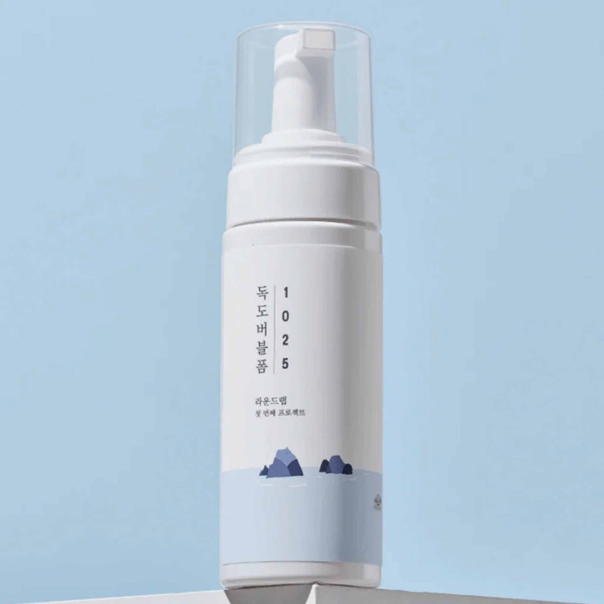 Schlichte weiße Flasche des Round Lab 1025 Dokdo Bubble Foam mit Pumpspender und blauem Insel-Design, vor hellblauem Hintergrund stilvoll in Szene gesetzt.