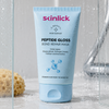 Eine blaue Tube der „Skinlick Peptide Gloss Bond Repair Mask“ liegt auf einer nassen Oberfläche mit Wassertropfen, neben einem Schwamm und einem Glas.