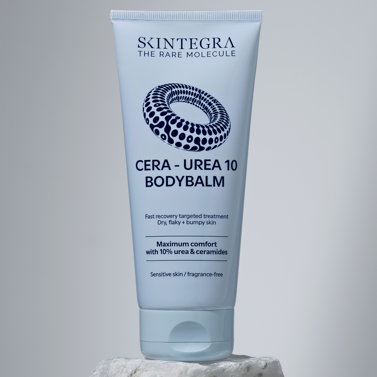 Eine Tube „Skintegra Cera-Urea 10 Bodybalm“ steht aufrecht auf einem Stein vor einem neutralen Hintergrund.