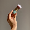 Eine Hand hält den SWEED Foundation Brush mit einem gold- und mintgrünen Griff vor einem beigen Hintergrund.
