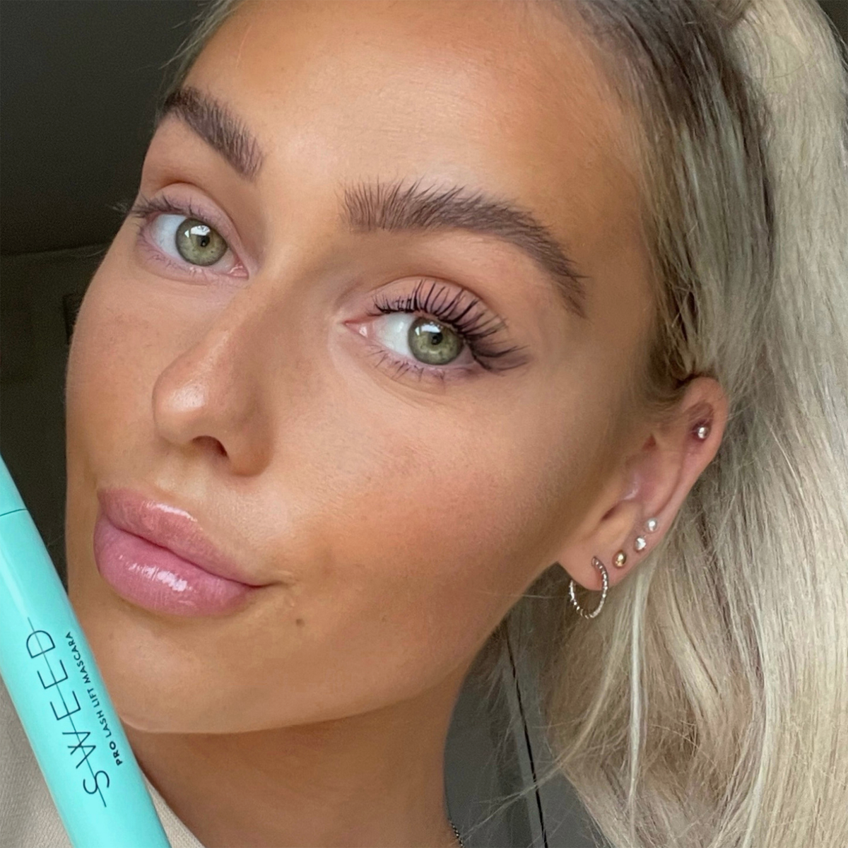 Eine Frau mit grünen Augen, deren Wimpern besonders lang wirken, hält die türkisfarbene Tube der SWEED Lash Lift Mascara nahe an ihr Gesicht.