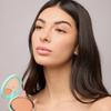 Eine Frau mit langen, dunklen Haaren hält die Kompaktdose „The Bronzing Powder“ von SWEED in der Hand und blickt mit ausdruckslosem Gesicht aus dem Bild.