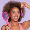 Frau mit lockigem Haar lächelt und hält eine große pink-lila UNbrush® Bürste sowie eine Mini-Version am Schlüsselanhänger hoch.