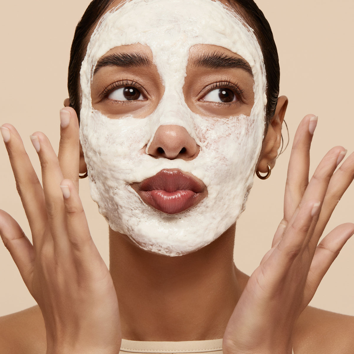 Eine Frau mit einer weißen Gesichtsmaske, die ein Schmollgesicht macht und ihre Hände in die Nähe ihrer Wangen hebt, präsentiert vor einem beigen Hintergrund Rice Enzyme Brightening Cleansing Powder.