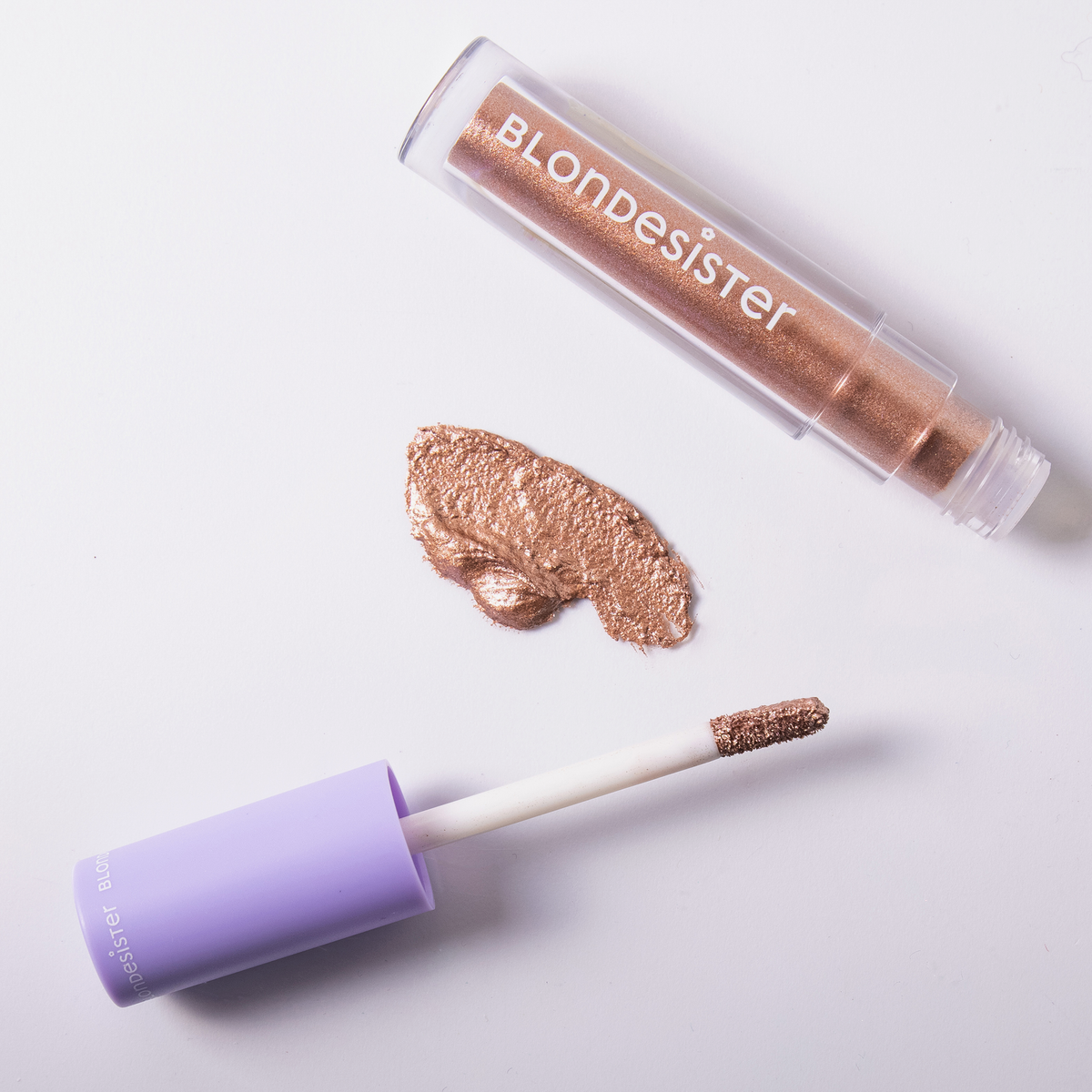 Eine Tube BLONDESISTER GLOWEYE EYESHADOW mit dem dazugehörigen Applikator und einem Farbmuster in Bronzeschimmer auf weißem Hintergrund.