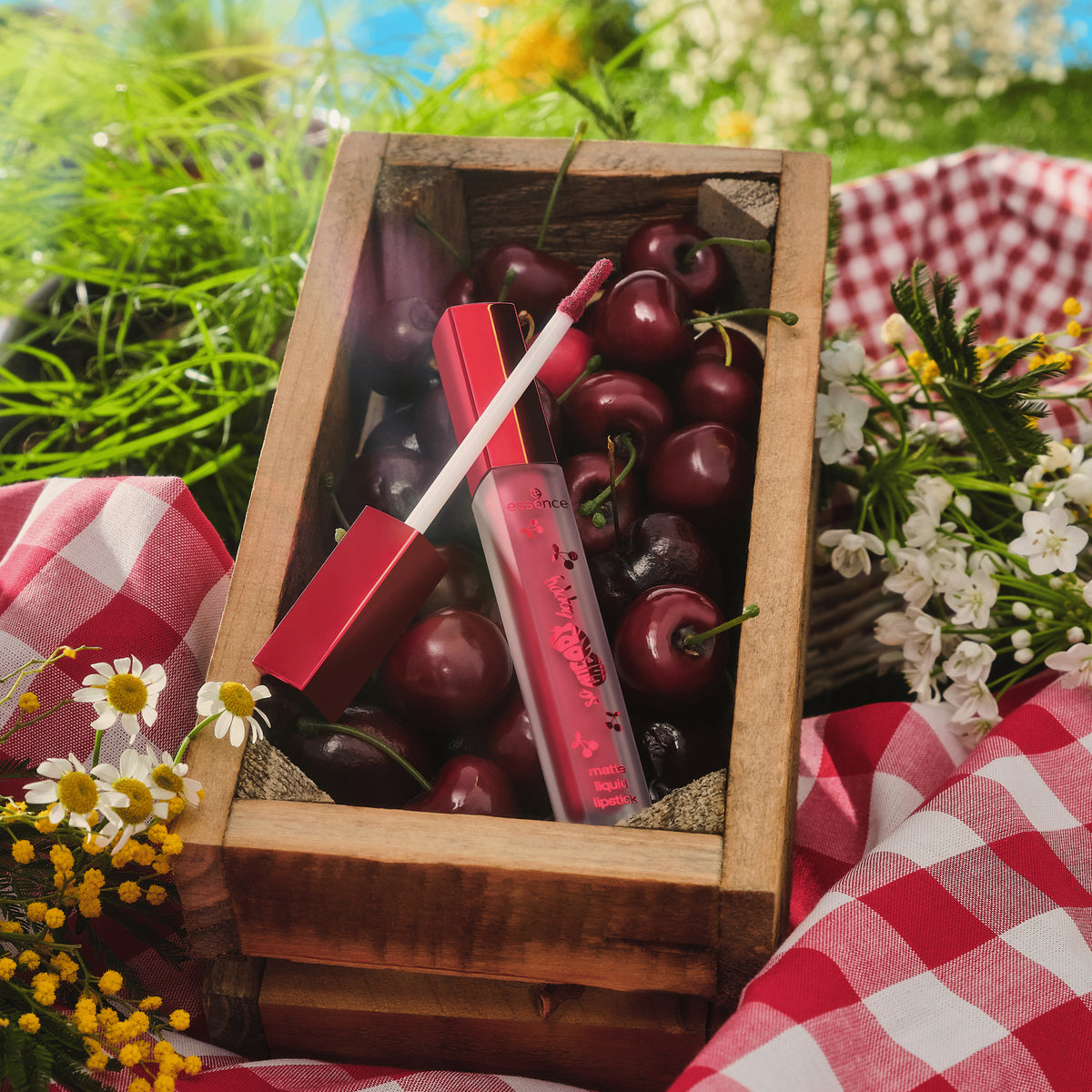 SO CHERRY Happy Matte Liquid Lipstick 01 mit Applikator, präsentiert auf frischen Kirschen in einer Holzbox, umgeben von Blumen und Grün.