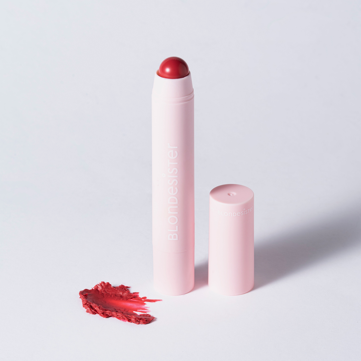 BLONDESISTER 2 IN 1 – DU HAST DIE WAHL: LIPPEN ODER WANGEN, rosa Tube ohne Verschluss, aus der ein roter Stift herausragt, daneben ein Fleck auf weißem Untergrund.