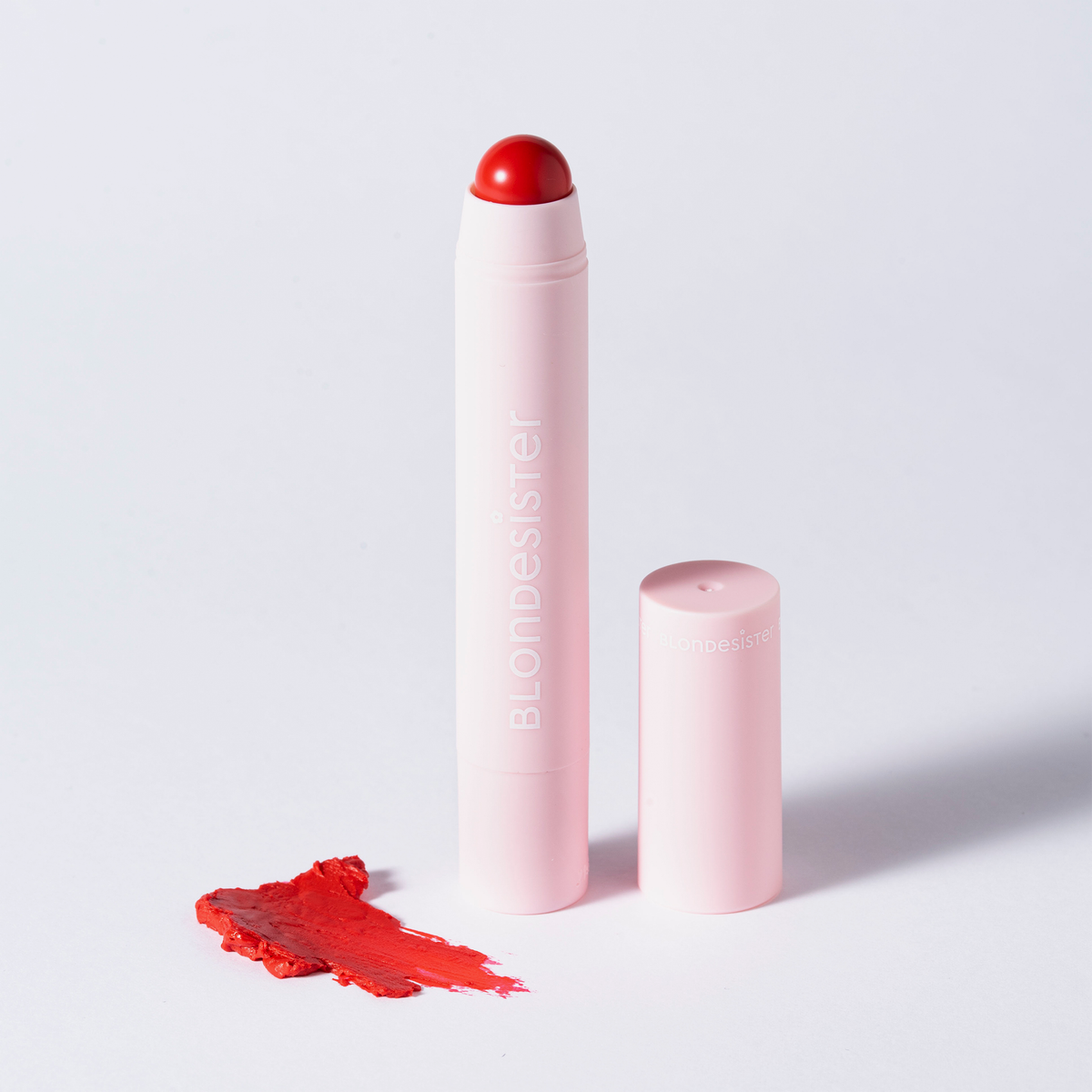 Eine „BLONDESISTER 2 IN 1 – IT’S UP TO YOU LIP OR CHEEK“-Tube steht mit abgenommener Kappe neben einem roten Fleck auf einer weißen Oberfläche.
