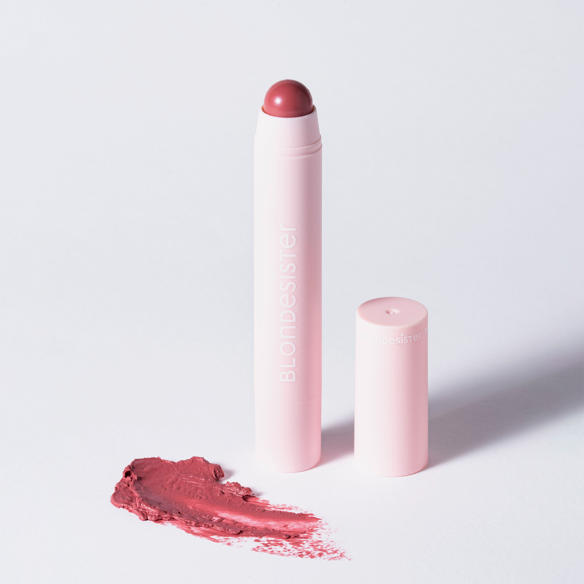 Ein „BLONDESISTER 2 IN 1 – IT’S UP TO YOU LIP OR CHEEK“-Stift, ohne Kappe, neben einem rosafarbenen Fleck auf einer weißen Oberfläche.