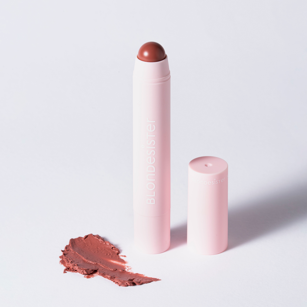 Ein rosa BLONDESISTER 2-IN-1-Stift „IT’S UP TO YOU – LIP OR CHEEK“ steht aufrecht und ohne Kappe neben einem bräunlichen Fleck auf weißem Hintergrund.