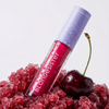 Eine Tube BLONDESISTER 2 IN 1 SWEET GLOSS TINTED LIP OIL steht auf rotem zerstoßenem Eis neben einer Kirsche; auf der Tube sind Wassertropfen zu sehen.