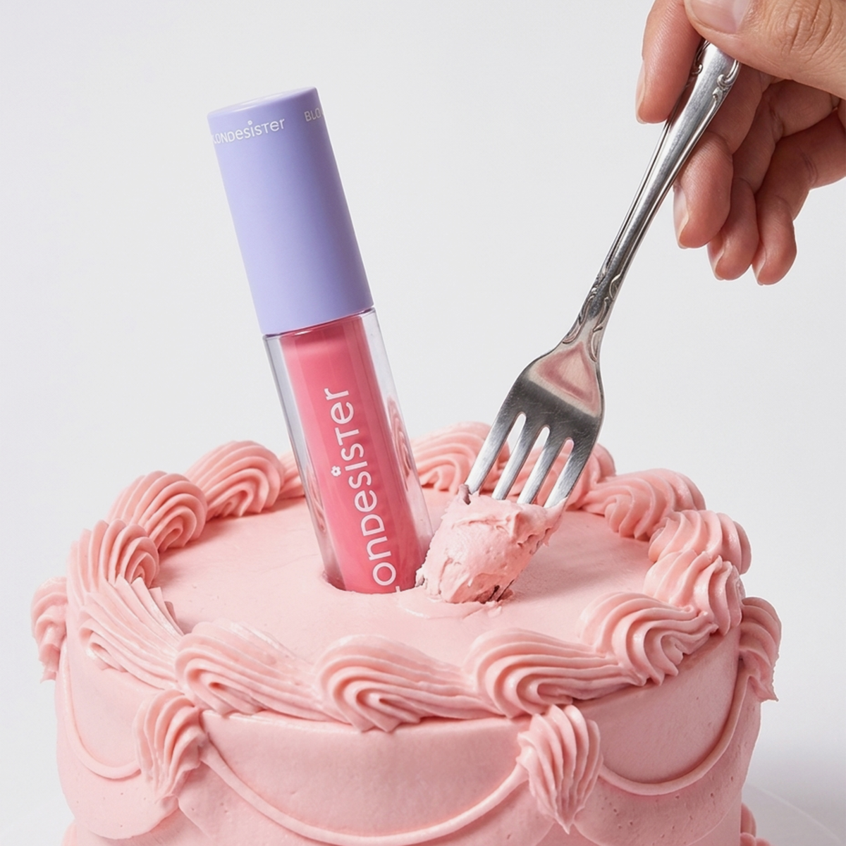 Eine Hand hält ein Stück rosa Kuchen, auf dem eine Tube „BLONDESISTER 2 IN 1 SWEET GLOSS TINTED LIP OIL“ aufrecht steht.