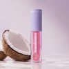 Ein rosa „BLONDESISTER 3 IN 1 CUTE LIPPY LIP OIL“ steht aufrecht neben einer aufgebrochenen Kokosnuss auf einer glänzenden Oberfläche.