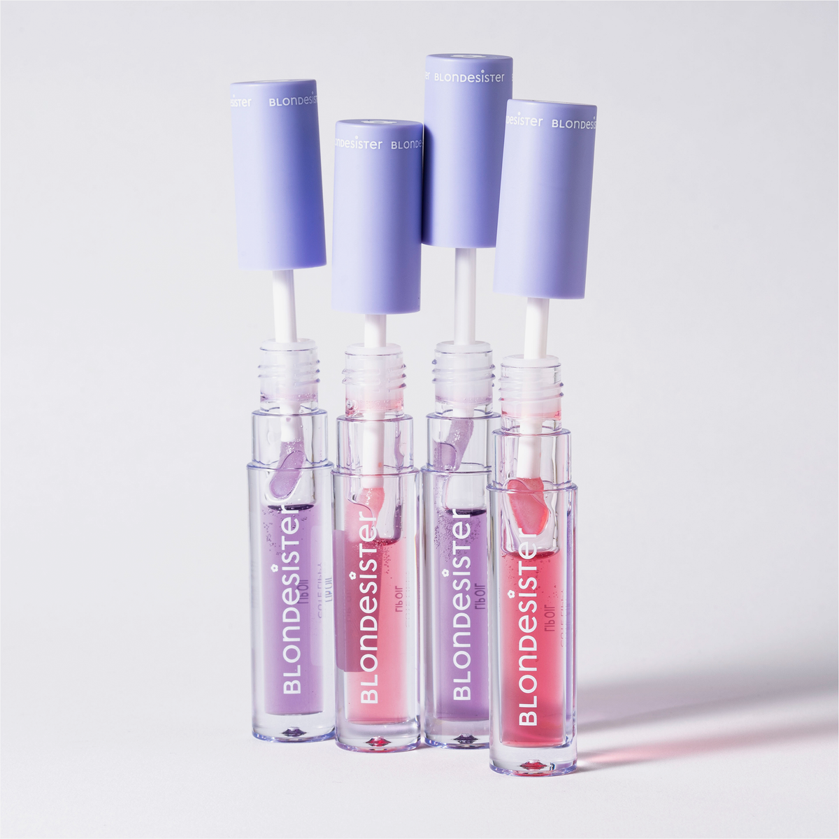 Vier aufrecht stehende Tuben des Produkts „BLONDESISTER 3 IN 1 CUTE LIPPY LIP OIL“ mit violetten Verschlusskappen zeigen im Inneren rosa und violette Farbtöne.