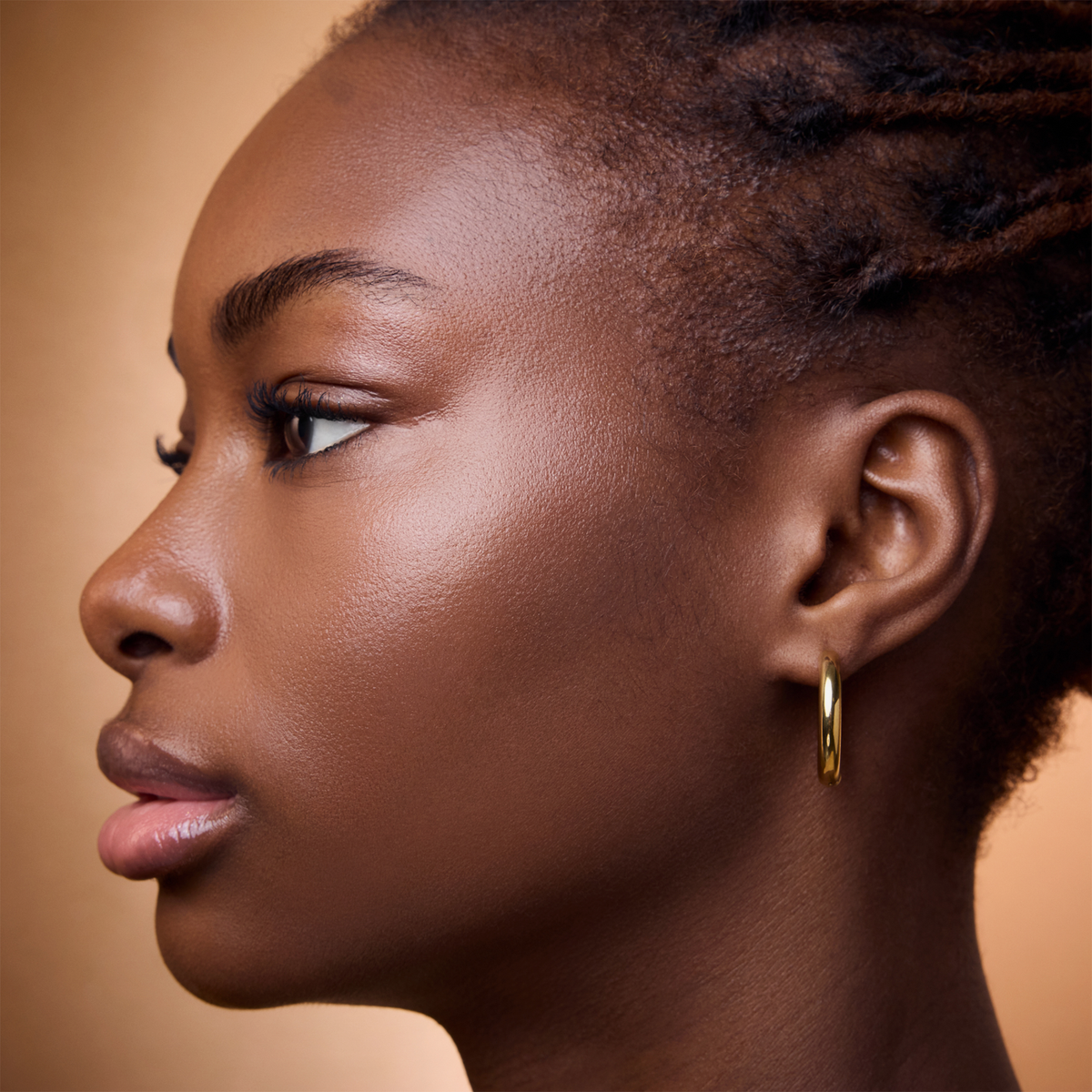 Eine schwarze Frau mit Braids und Creolen präsentiert vor einem sanften beigen Hintergrund den Wonder Woman Butter Bronzer Stick.