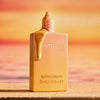 Eine geöffnete Flasche Catrice Cosmetics MIDNIGHT SUN Glow Drops mit goldener Flüssigkeit, die vor einem Strandhintergrund mit Sonnenuntergang tropft.