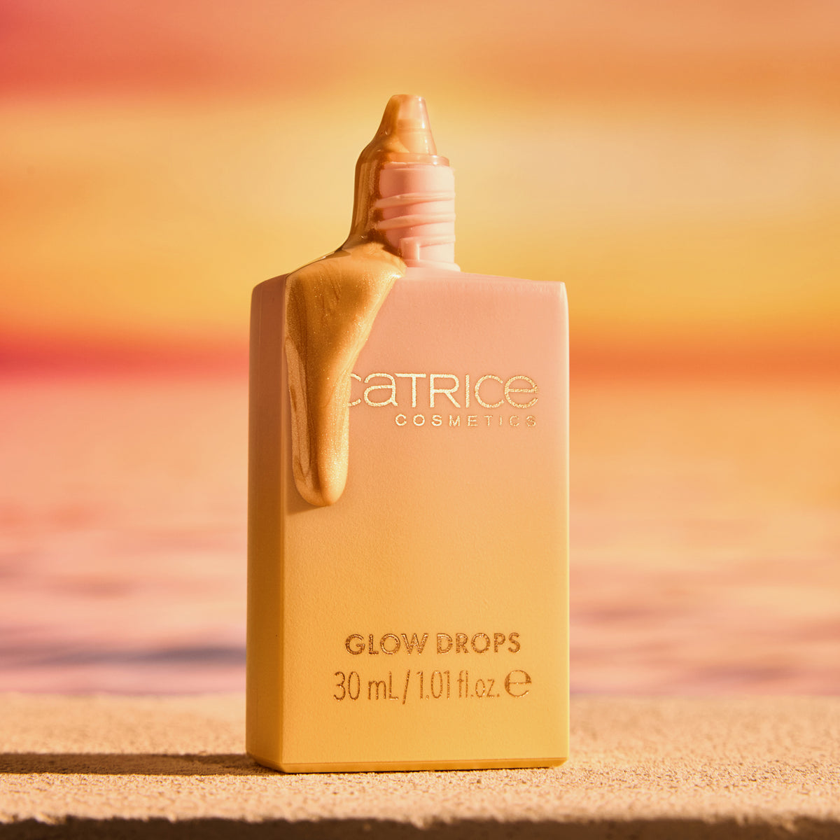 Eine geöffnete Flasche Catrice Cosmetics MIDNIGHT SUN Glow Drops mit goldener Flüssigkeit, die vor einem Strandhintergrund mit Sonnenuntergang tropft.